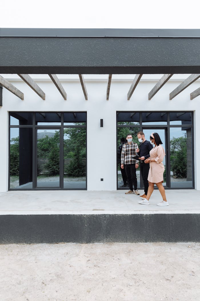 Three real estate agents wearing masks showcase a sleek, modern home exterior to clients.